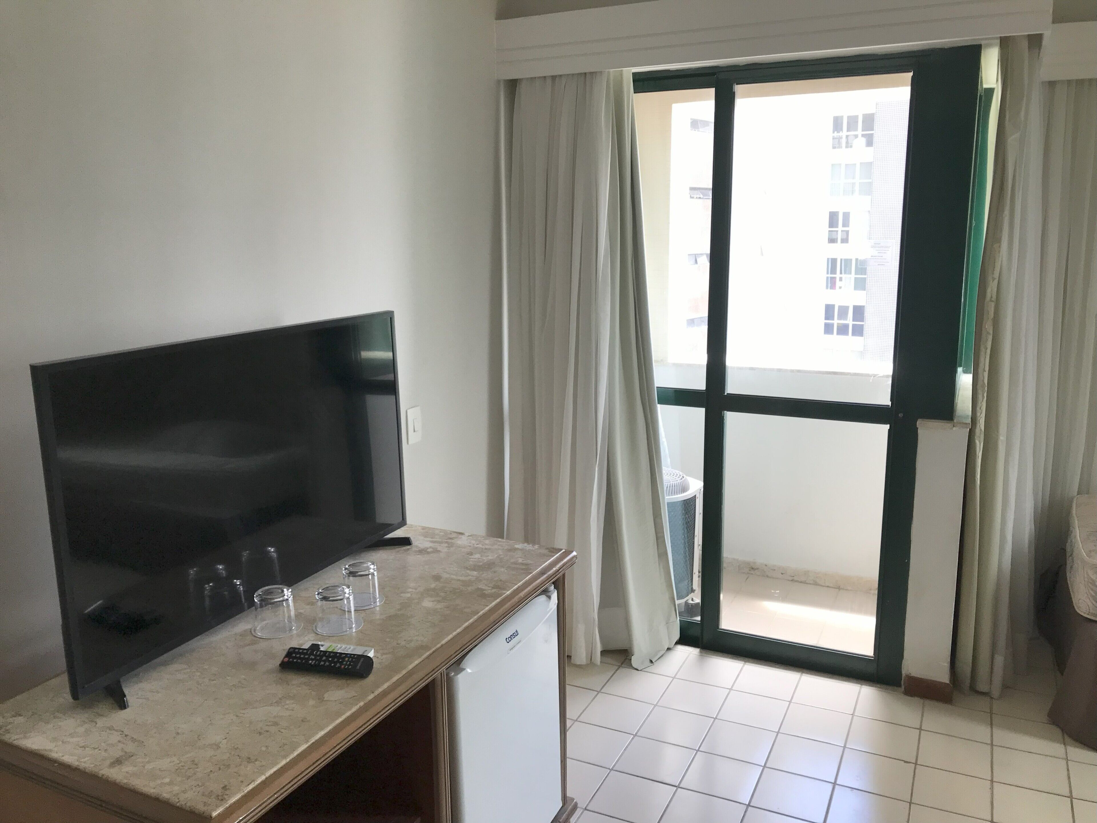 Una habitación con televisión de pantalla plana, un refrigerador pequeño, cafetera y puerta corrediza que da a un balcón.