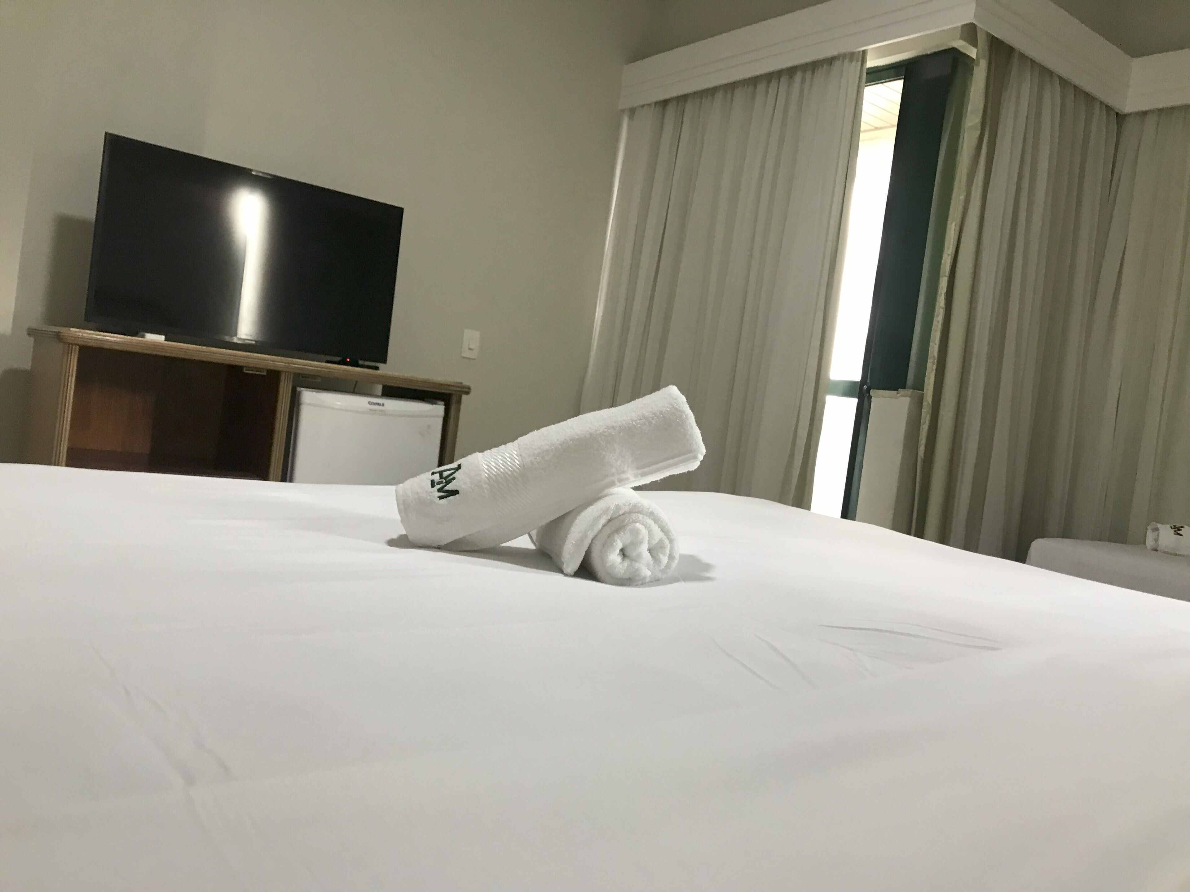 Een hotelkamer met een bed, een televisie op een standaard en een raam met gordijnen.
