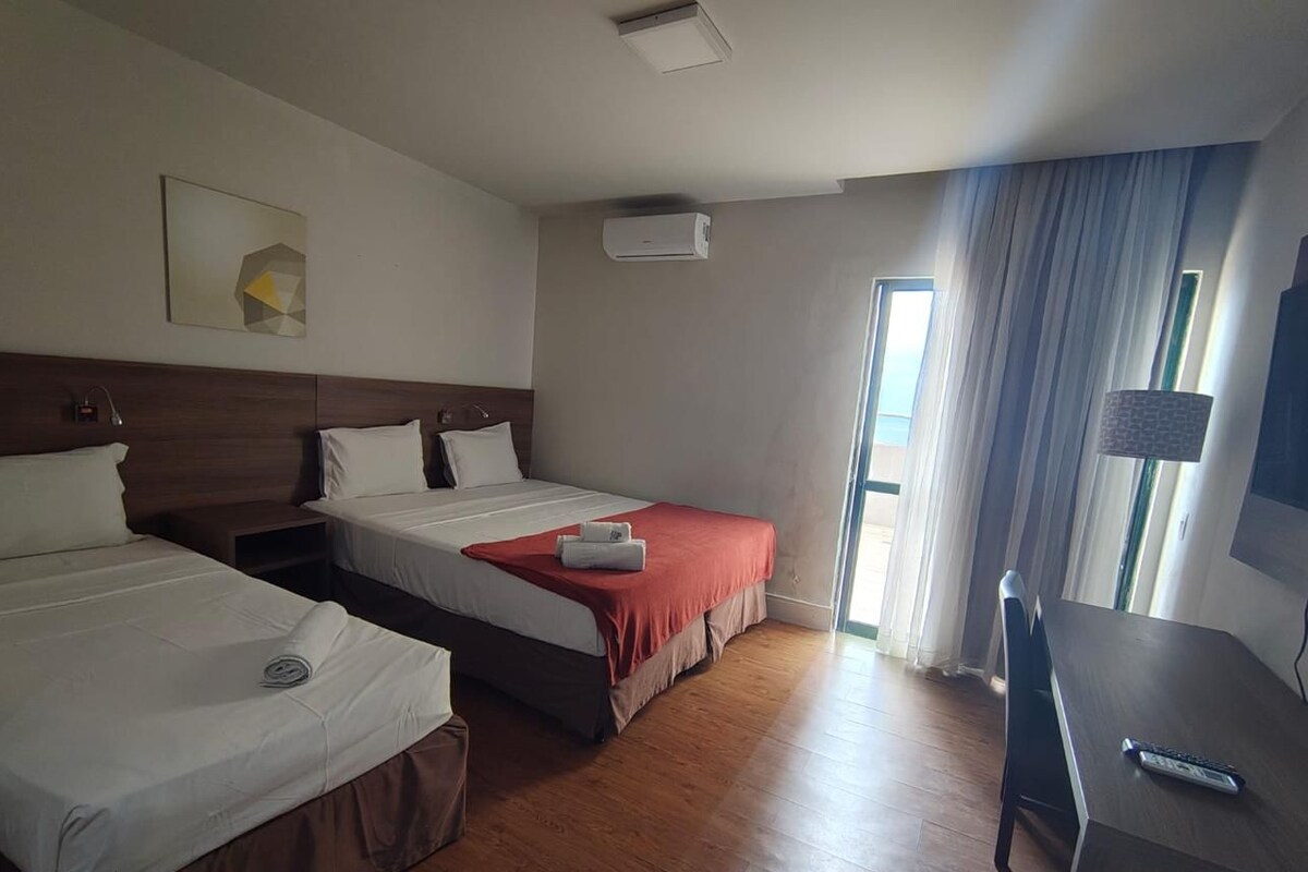 Quarto de hotel com duas camas, uma escrivaninha e uma janela com cortinas.