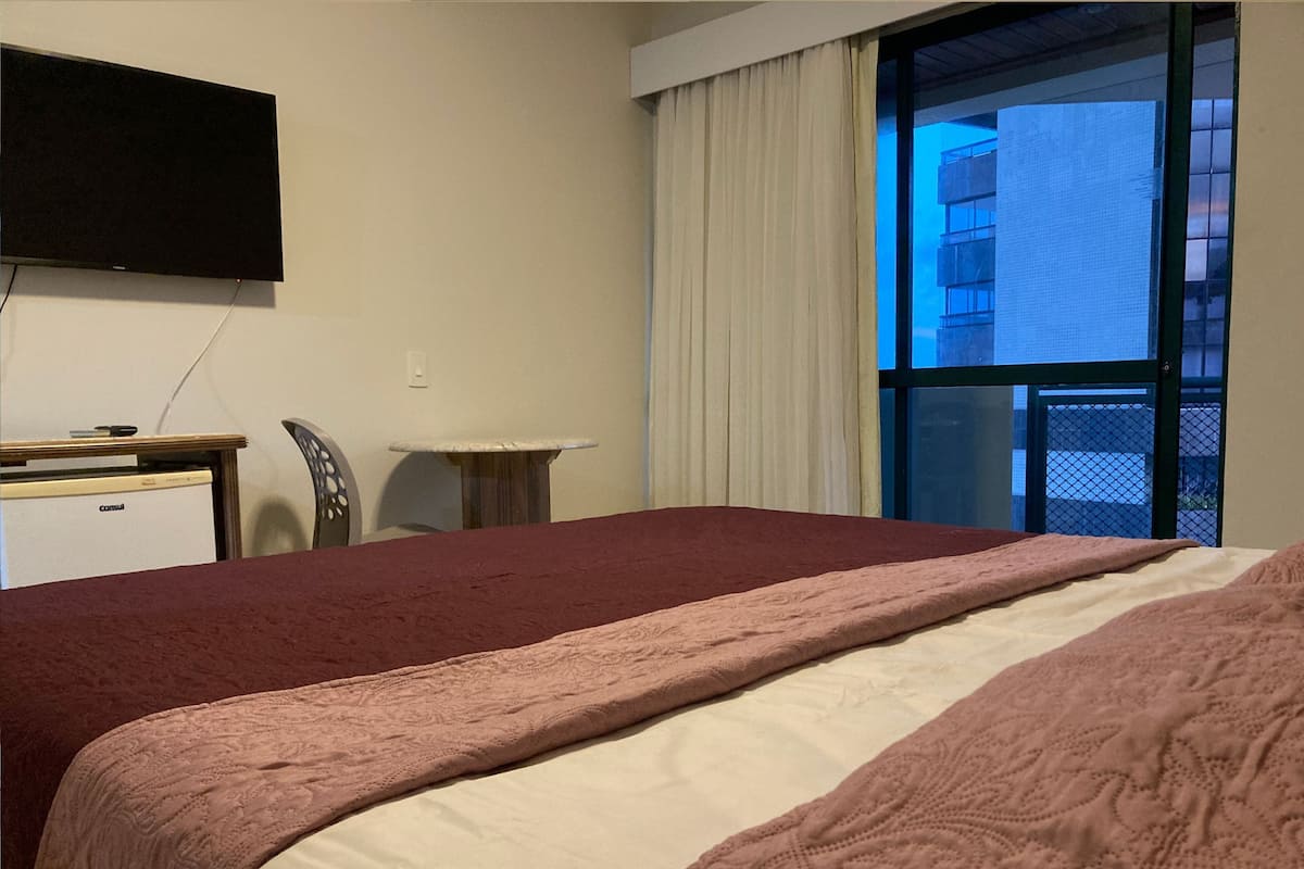 A hotel room with a bed, a desk, a chair, a television, and a window with a view of a building.