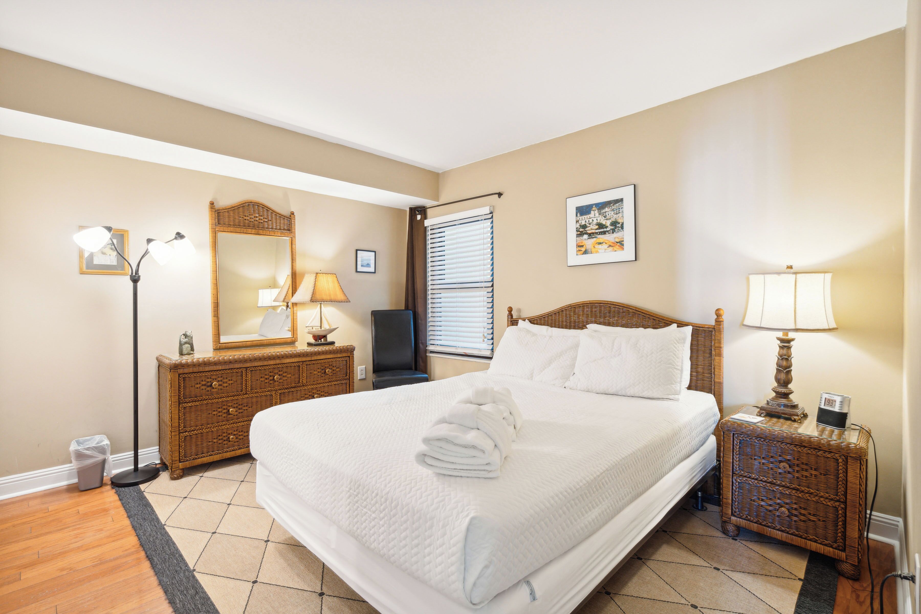A hotel room with a large bed, a wooden dresser, a nightstand with a lamp, a mirror, and a window with blinds.