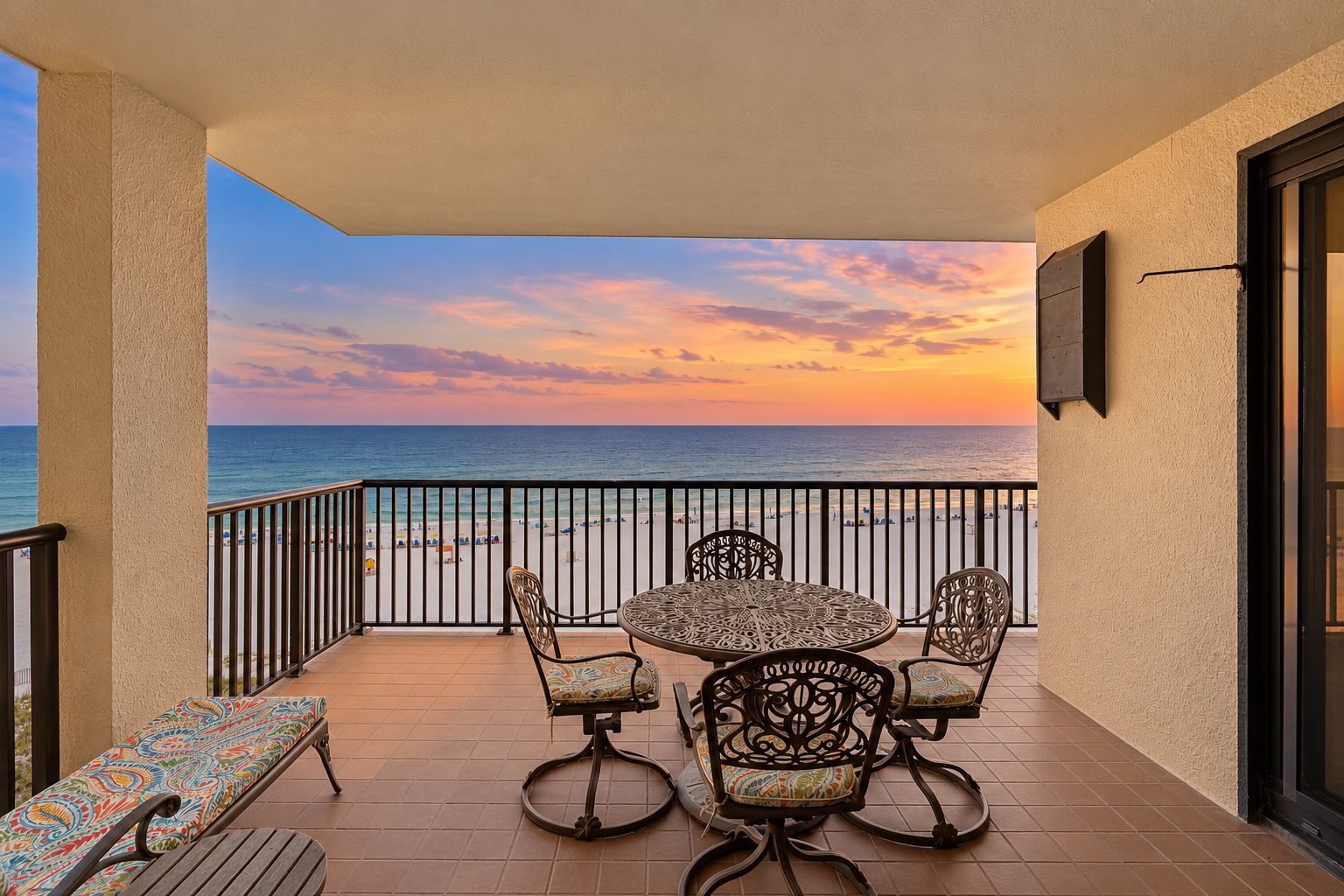 Un balcon avec une table et des chaises, donnant sur une plage et l’océan au coucher du soleil.