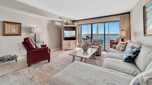 Amplia sala de estar con un gran ventanal que ofrece vista al océano, un sillón de cuero rojo, un sofá gris y una mesa de centro de madera.