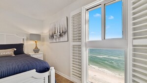 Un dormitorio con cama, mesita de noche, lámpara, un cuadro y ventana con vista a la playa.