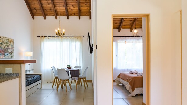 A room with a bed, a dining table with chairs, a chandelier, and a TV. - Luna Azul. Condo. Fast Wifi. Free Parking. (San Carlos de Bariloche)