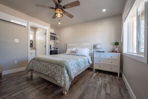 Um quarto com cama, mesa de cabeceira, ventoinha no teto, janela e porta que dá para outro cômodo.