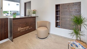 Un área de recepción con un escritorio de madera, un sillón color beige, un espejo grande y un estante de madera montado en la pared.