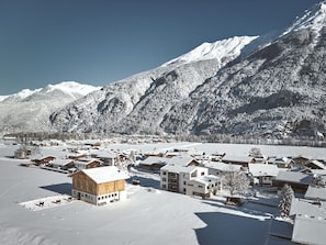 Un village enneigé avec des maisons traditionnelles en bois, un grand bâtiment à la façade en bois, et des montagnes enneigées en arrière-plan.