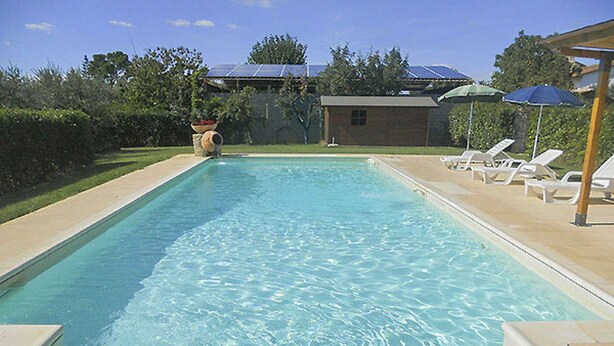 Una piscina rectangular con agua azul cristalina, rodeada por una solería con baldosas. Hay sillas de descanso y sombrillas a la derecha, y un edificio con techo de paneles solares al fondo.