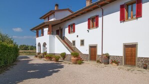 Un edificio de dos pisos con fachada blanca, contraventanas rojas y detalles de piedra. Tiene varias puertas y ventanas, y el suelo está cubierto de grava.