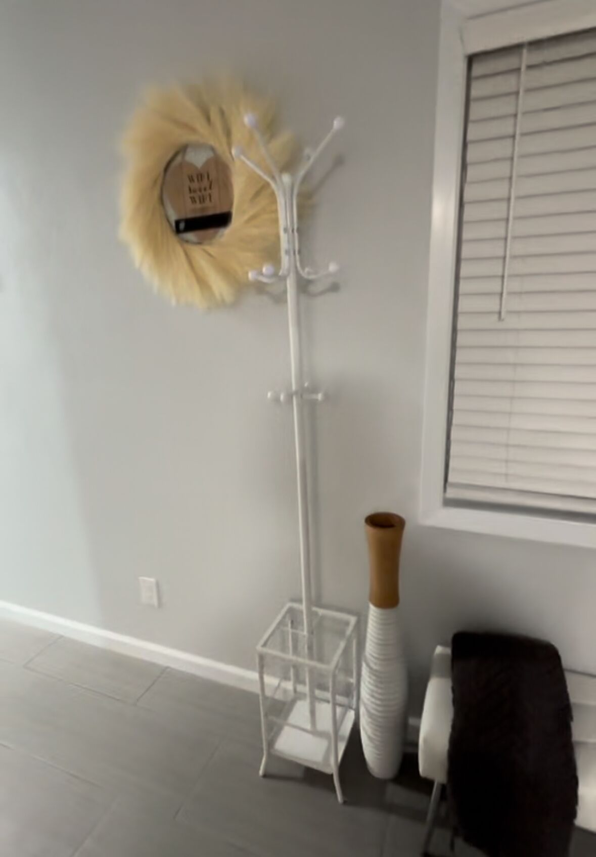 Una habitación moderna con suelo blanco, pared blanca, ventana con persianas y un perchero blanco decorado con un detalle de textura suave, junto a un jarrón blanco alto.