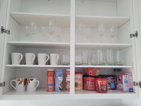 A kitchen cabinet with shelves containing various items including mugs, wine glasses, and coffee products.