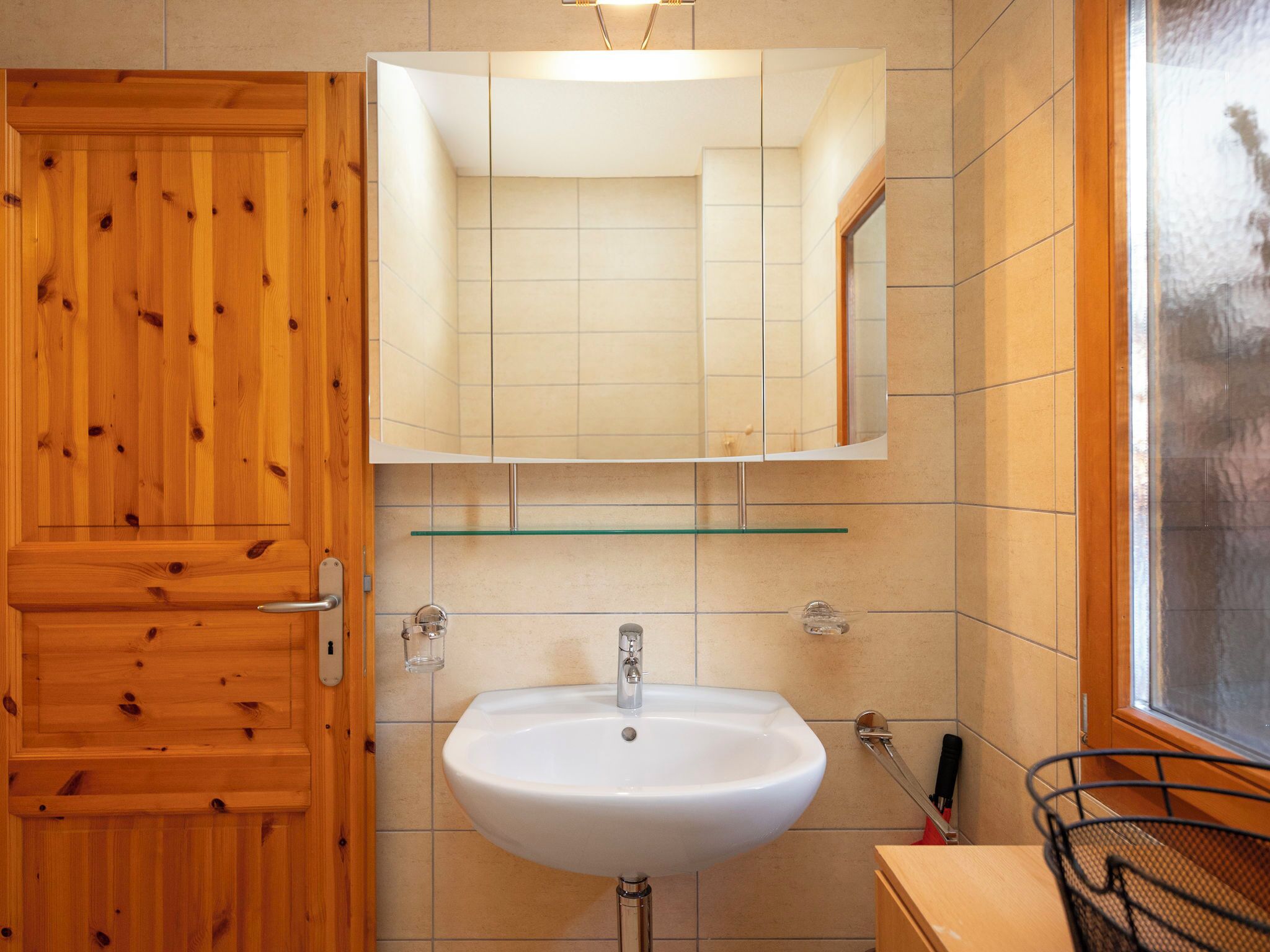 A bathroom with a wooden door, a white sink, a mirror, and a window.