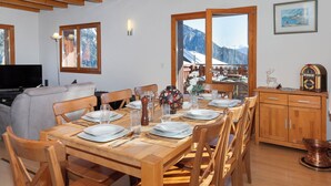 A dining area with wooden furniture, a table set for four, and a view of mountains through large windows. - Orchidée by Interhome (Ovronnaz)
