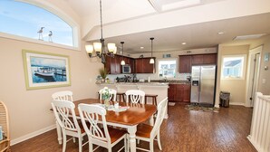 A dining area with a wooden table and white chairs, a kitchen with wooden cabinets and stainless steel appliances, and a view of the outside through a large window. - Oceanside, Hot Tub, Community Pool, CB51, OBXcape (Corolla)