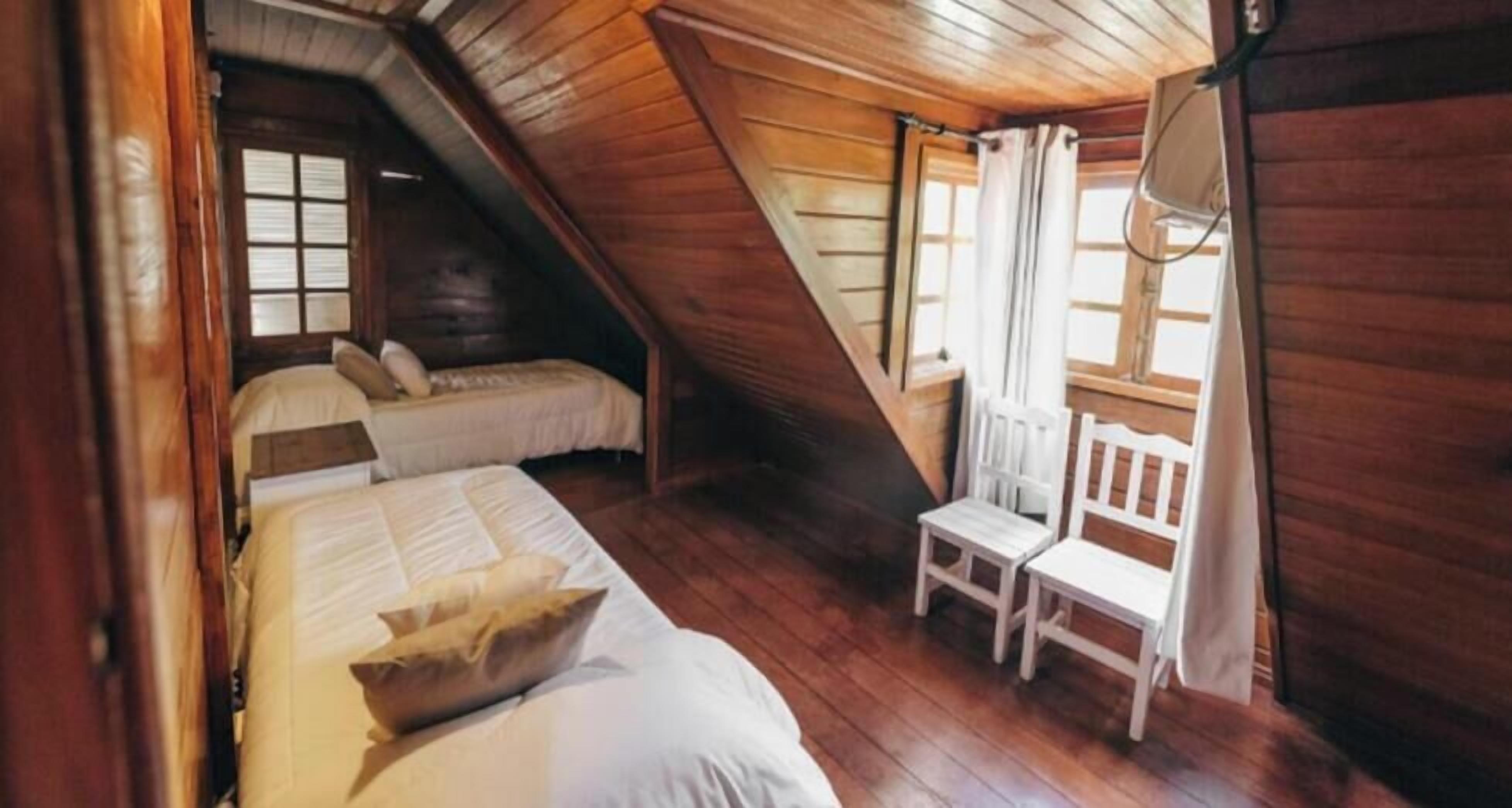 A room with wooden walls and sloped ceilings, two beds, a chair, and a window with curtains.