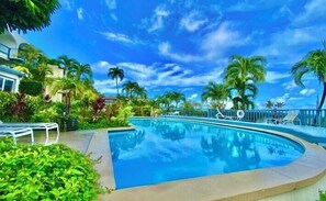 Une piscine entourée de palmiers et de verdure, avec des chaises longues et un ciel d’un bleu limpide.