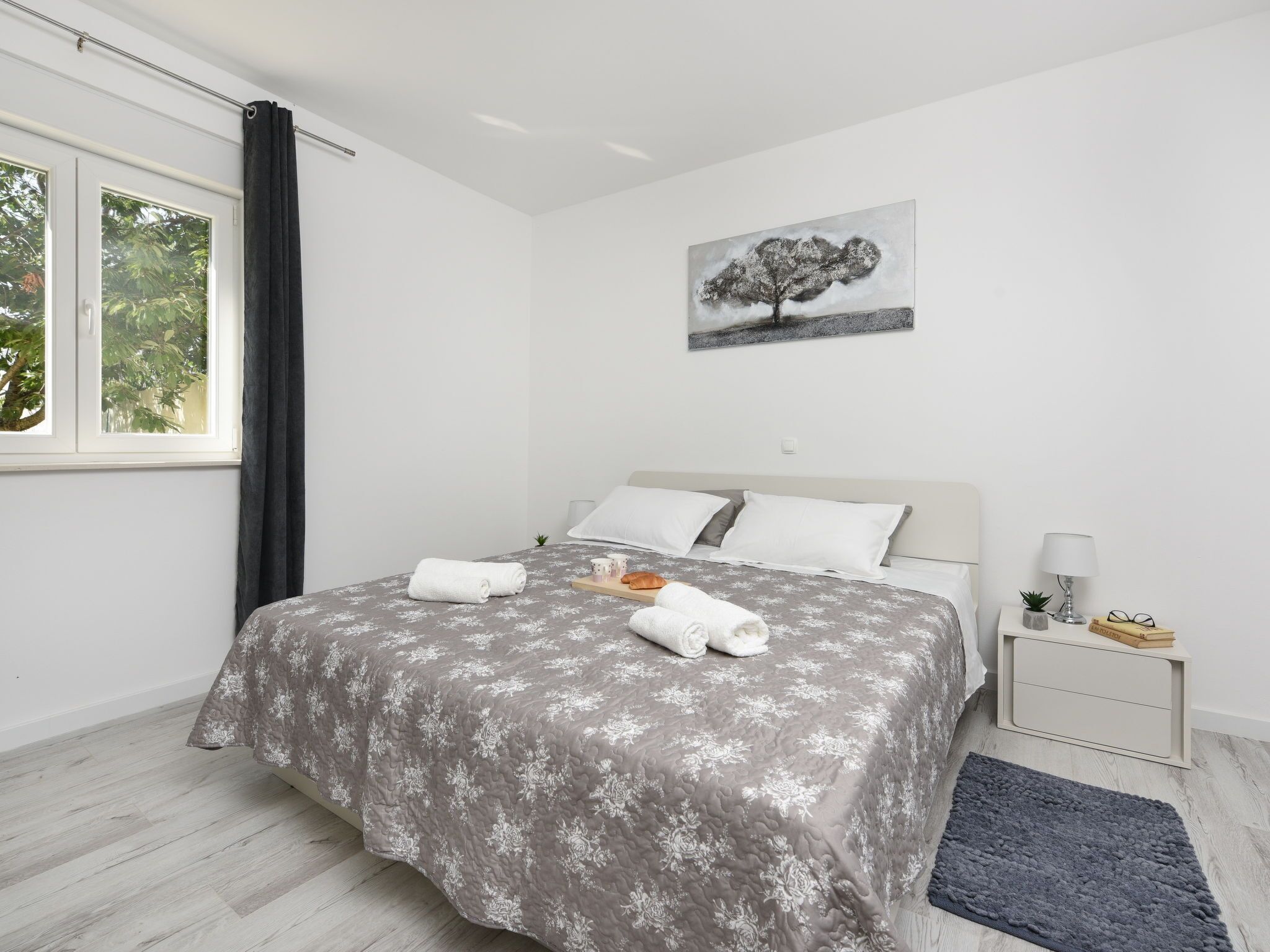 Un dormitorio con cama, mesita de noche, lámpara, un cuadro y ventana con cortinas.