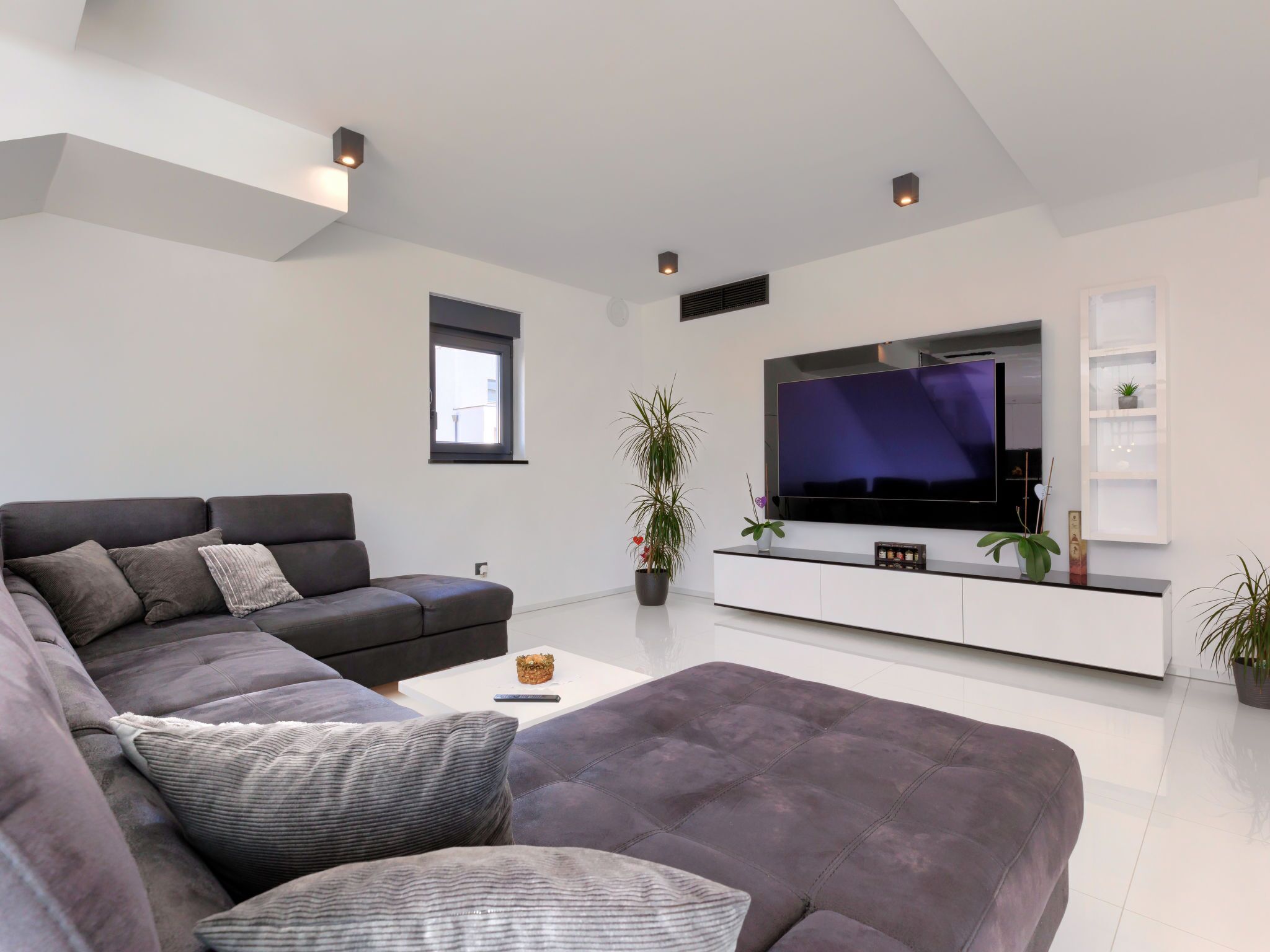 A modern living room with a large flat-screen TV, a dark grey sectional sofa, a white coffee table, and a minimalist white entertainment unit.