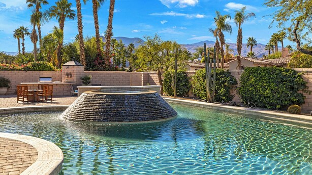 Una piscina con una cascada escalonada, rodeada de palmeras y un muro de ladrillo.