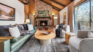 Ein gemütliches Wohnzimmer mit Kamin, grünen und grauen Sofas, einem runden Holztisch und einem großen Fenster mit Vorhängen.