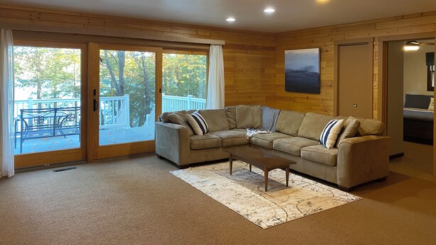 A cozy living room with a sectional sofa, a coffee table, and a large window with a view of a deck and trees. - Lake Lucerne Family Getaway, private setting close to town (Crandon)