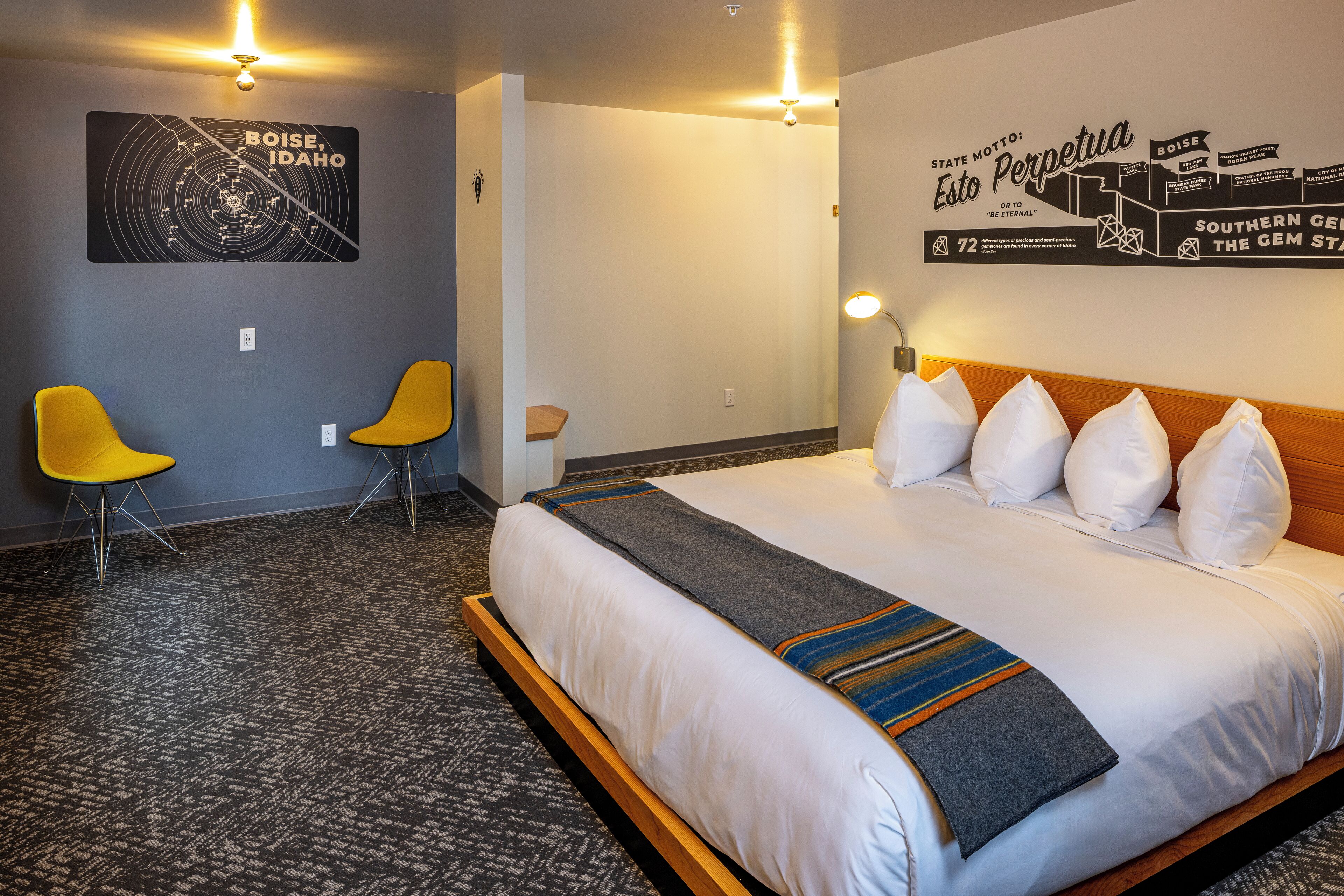 A hotel room with a large bed, two yellow chairs, a wall map of Boise, Idaho, and a wall mural with the motto "Esto Perpetua".