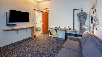 A modern hotel room with a flat-screen TV, a sofa, a desk with a chair, and a mirror.