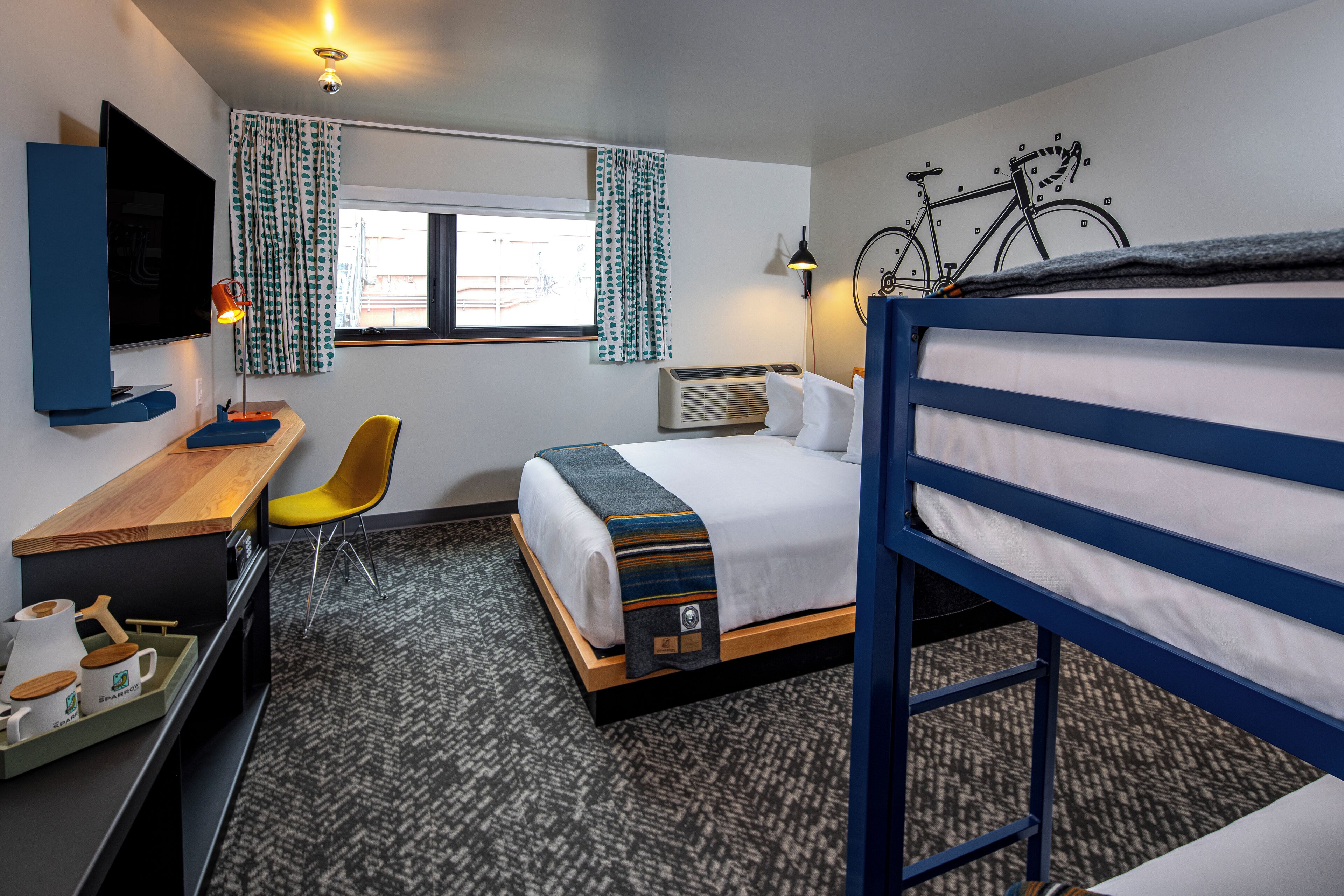 A hotel room with a bunk bed, a desk with a chair, a TV, a window with curtains, and a bicycle mounted on the wall.