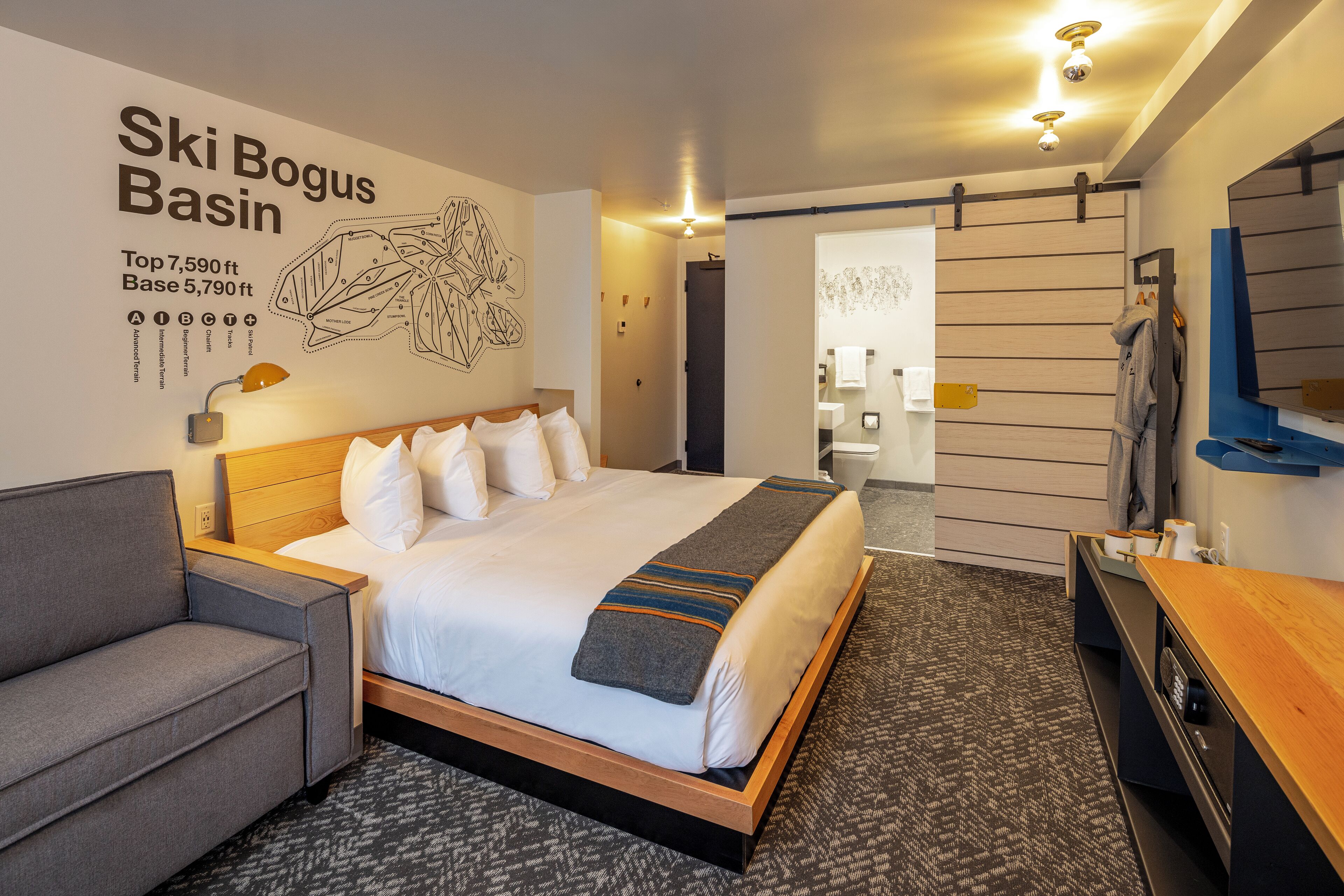 A hotel room with a large bed, a gray sofa, a wooden desk, and a wall with a ski-themed artwork.