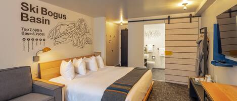 A hotel room with a large bed, a gray sofa, a wooden desk, and a wall with a ski-themed artwork.
