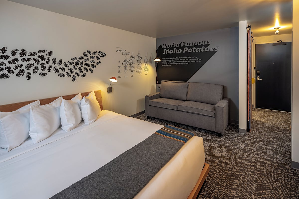 Quarto de hotel com uma cama grande, um sofá cinza, uma parede com diagrama de planta de batata e uma porta.
