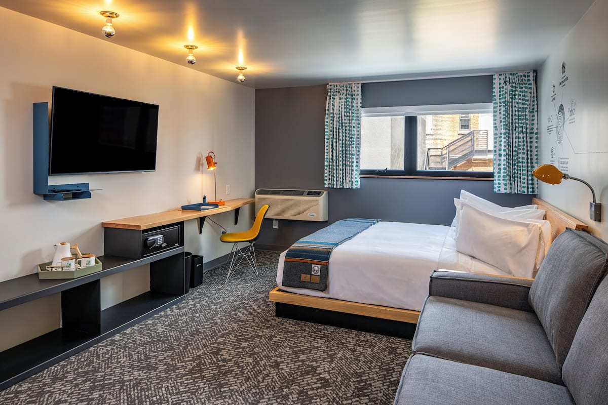 Quarto de hotel moderno com uma cama grande, uma escrivaninha com cadeira, televisão de tela plana e janela com cortinas.