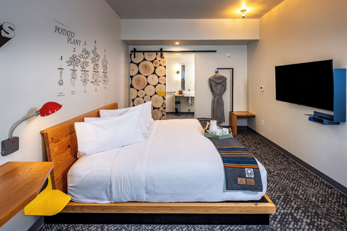 Quarto de hotel com cama de madeira, televisão de tela plana, banheiro com espelho e porta-toalhas fixado na parede.