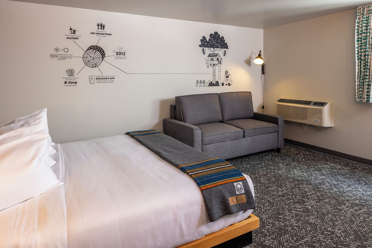 Quarto de hotel com uma cama, um sofá cinza, uma parede com diagrama circular, um abajur e um ar condicionado.
