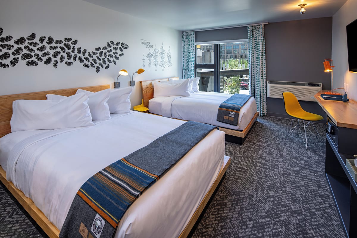 Quarto de hotel com duas camas, uma escrivaninha, uma cadeira e uma grande janela com vista para edifícios.