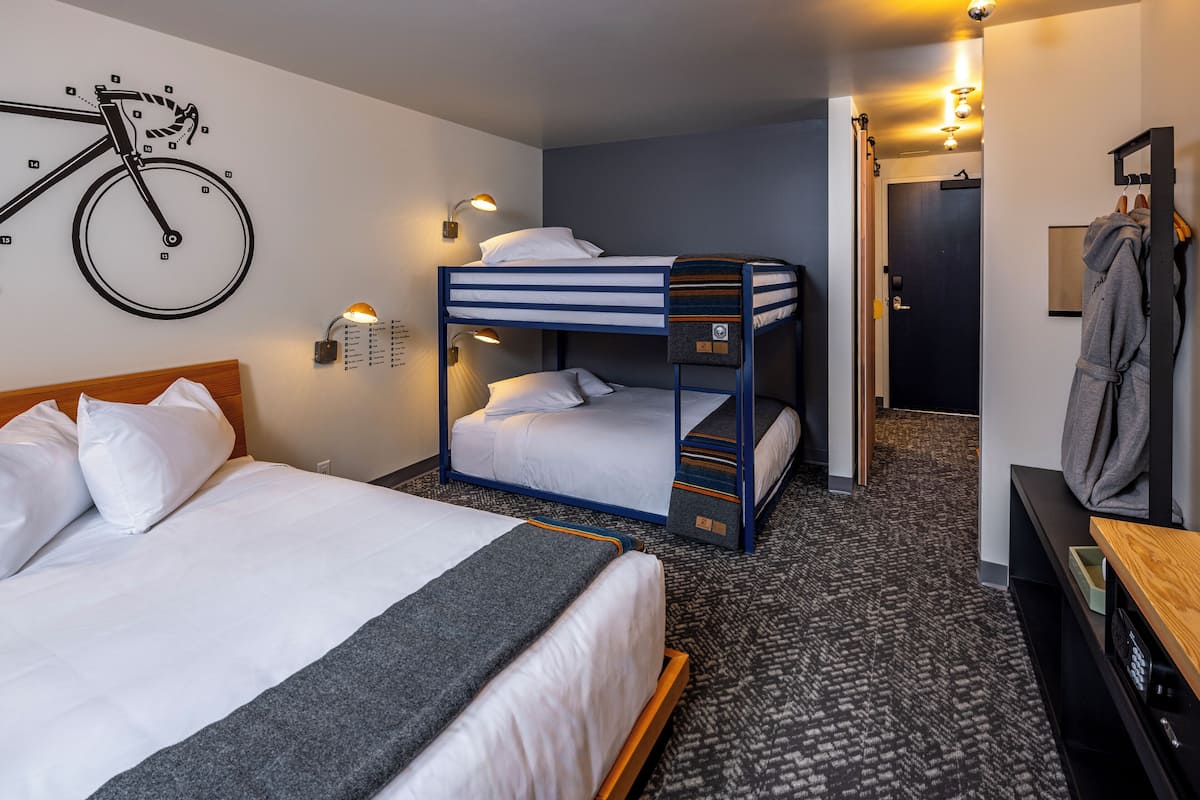 Quarto de hotel com uma cama de beliche, uma cama de solteiro, ilustração de uma bicicleta na parede, um porta-roupas e uma escrivaninha de madeira.