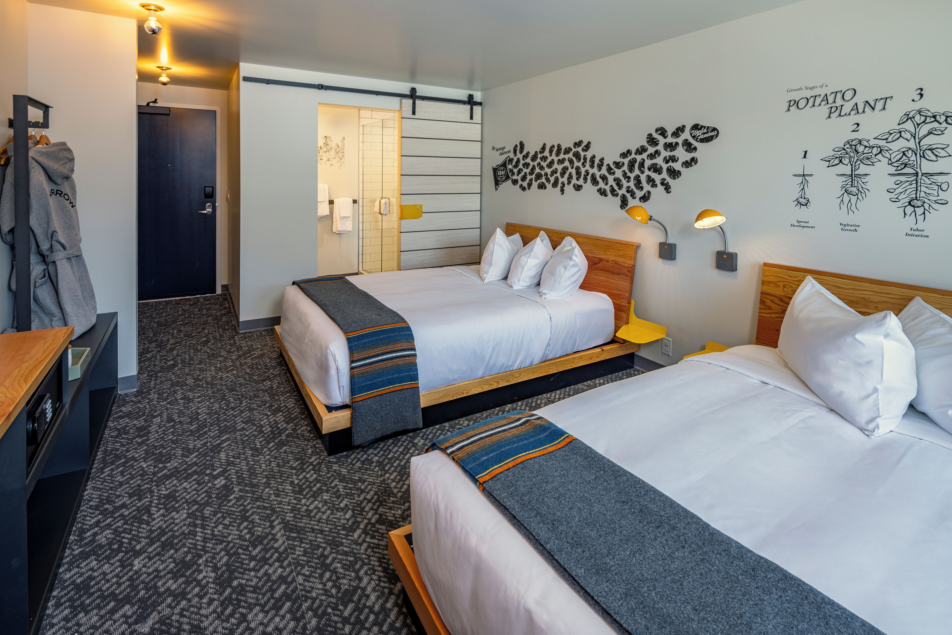A hotel room with two beds, a wooden headboard, a wall art of a potato plant, and a coat rack with a jacket.