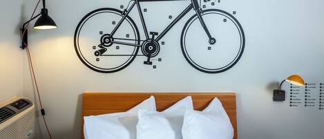 Ein Bett mit weißer Bettwäsche und Kissen, eine Wandillustration eines Fahrrads mit Beschriftung der Teile, eine Wandleuchte und ein Thermostat.