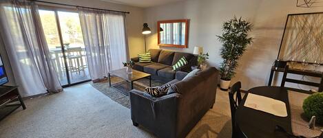 A living room with a sofa, coffee table, and a dining table with chairs. There is a TV on a stand, a potted plant, and a painting on the wall.