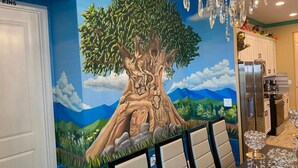Un coin repas agrémenté d’une fresque représentant un arbre, d’un lustre et d’une cuisine en arrière-plan.
