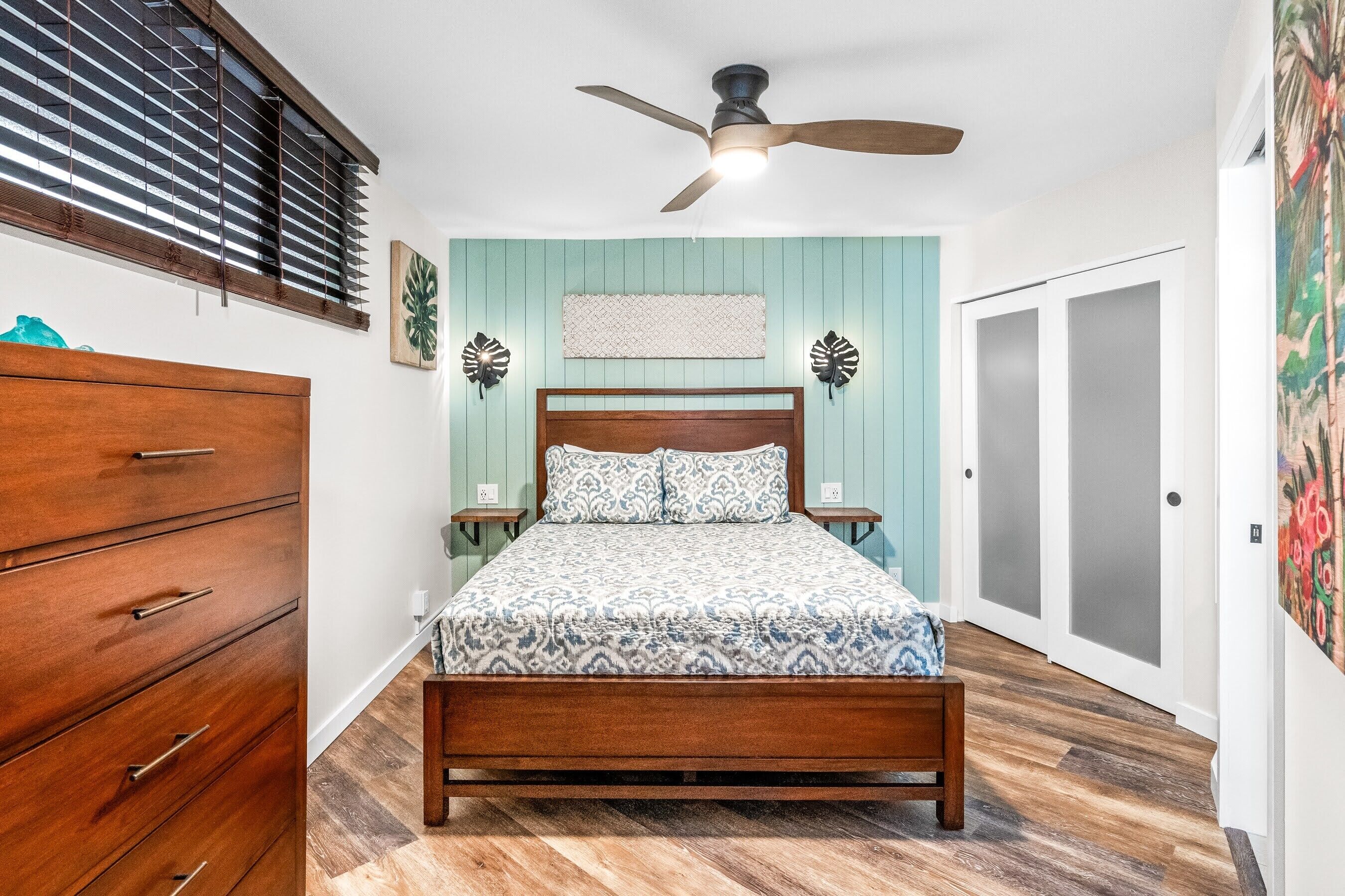 Une chambre à coucher avec un lit en bois, un ventilateur de plafond, une commode et une fenêtre avec des stores.