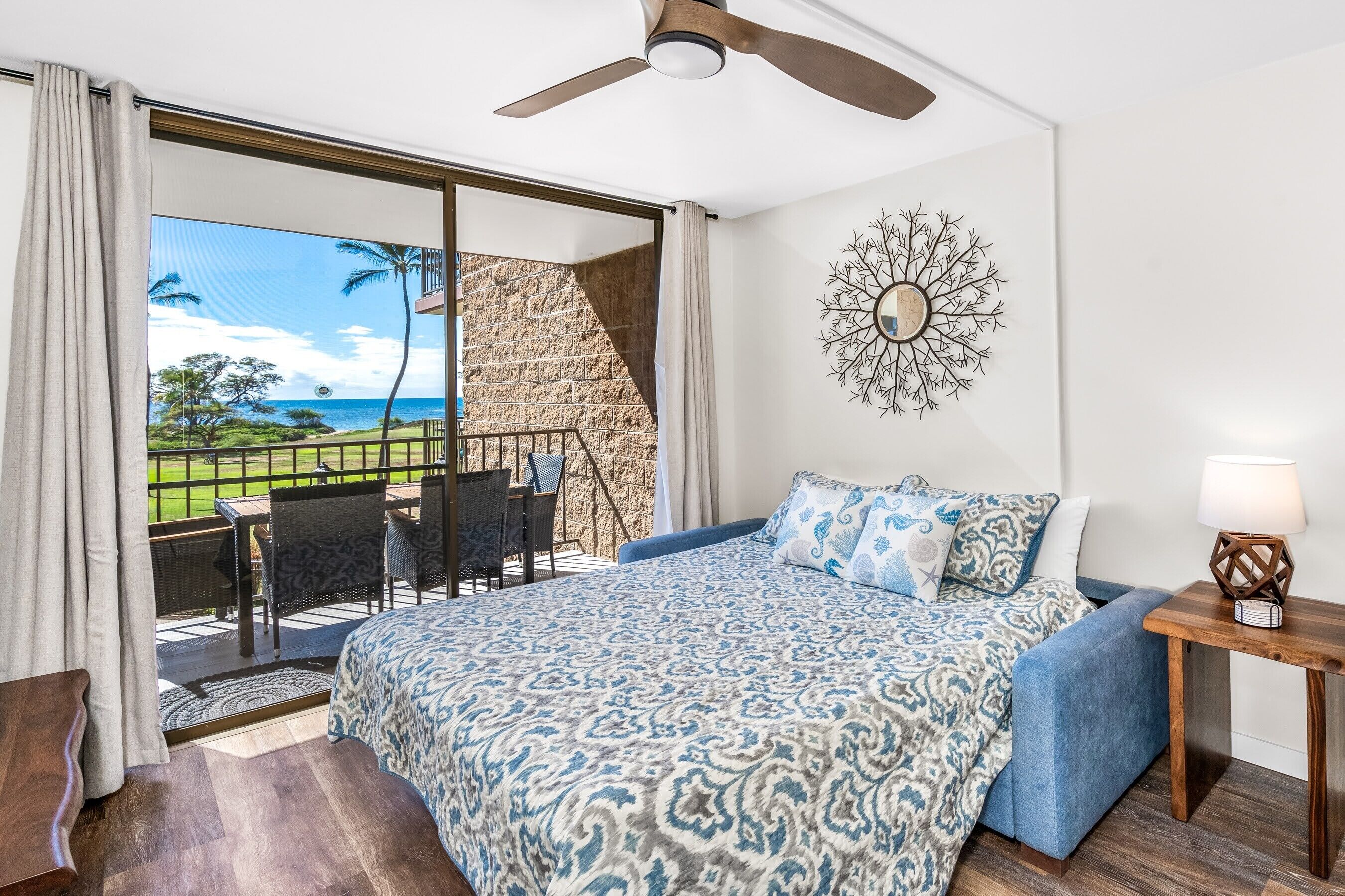 A bedroom with a large bed, a ceiling fan, a nightstand with a lamp, a wall clock, and a view of a balcony with outdoor furniture.