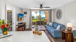 A living room with a blue sofa, a coffee table, and a TV on the wall. There is a balcony with a view of the ocean and palm trees. - 2 2 Oceanview Modern Resort Vistas (Kihei)