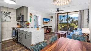 A modern kitchen with grey cabinets, a built-in oven, and a white countertop. There is a dining area with a wooden table and blue chairs. - 2 2 Oceanview Modern Resort Vistas (Kihei)