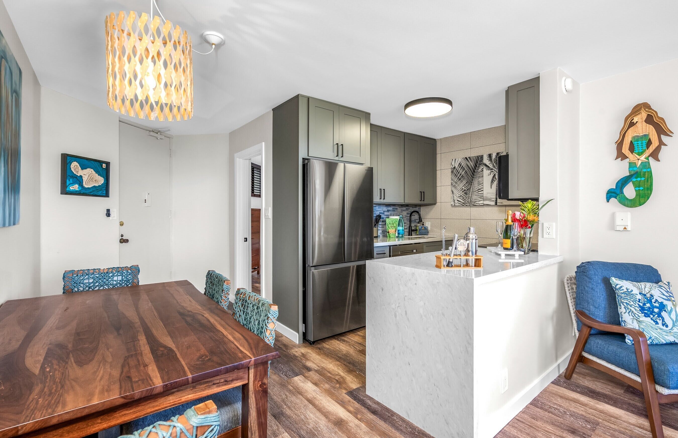 A modern kitchen with a stainless steel refrigerator, a dining area with a wooden table and patterned cushions, and a blue sofa with a mermaid decoration.