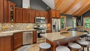 A kitchen with wooden cabinets, a large island with stools, stainless steel appliances, and a view of the outdoors through windows. - Modern, secluded luxury cabin near Castle Rock & Petenwell Lakes + Wisc. Dells (Necedah)