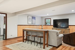 A modern living room with a wooden dining table, bar stools, a sofa, and a television.