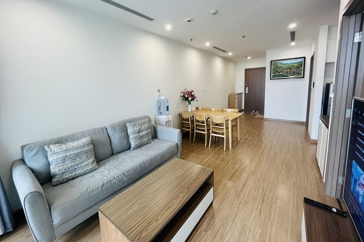 A modern living room with a grey sofa, wooden coffee table, dining table with chairs, and a TV on the right.