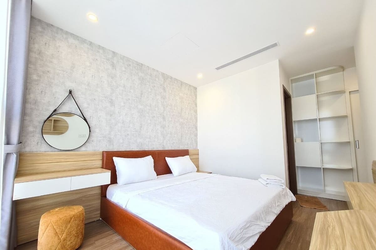 A modern hotel room with a wooden bed, a round mirror, a small stool, and a white wardrobe.